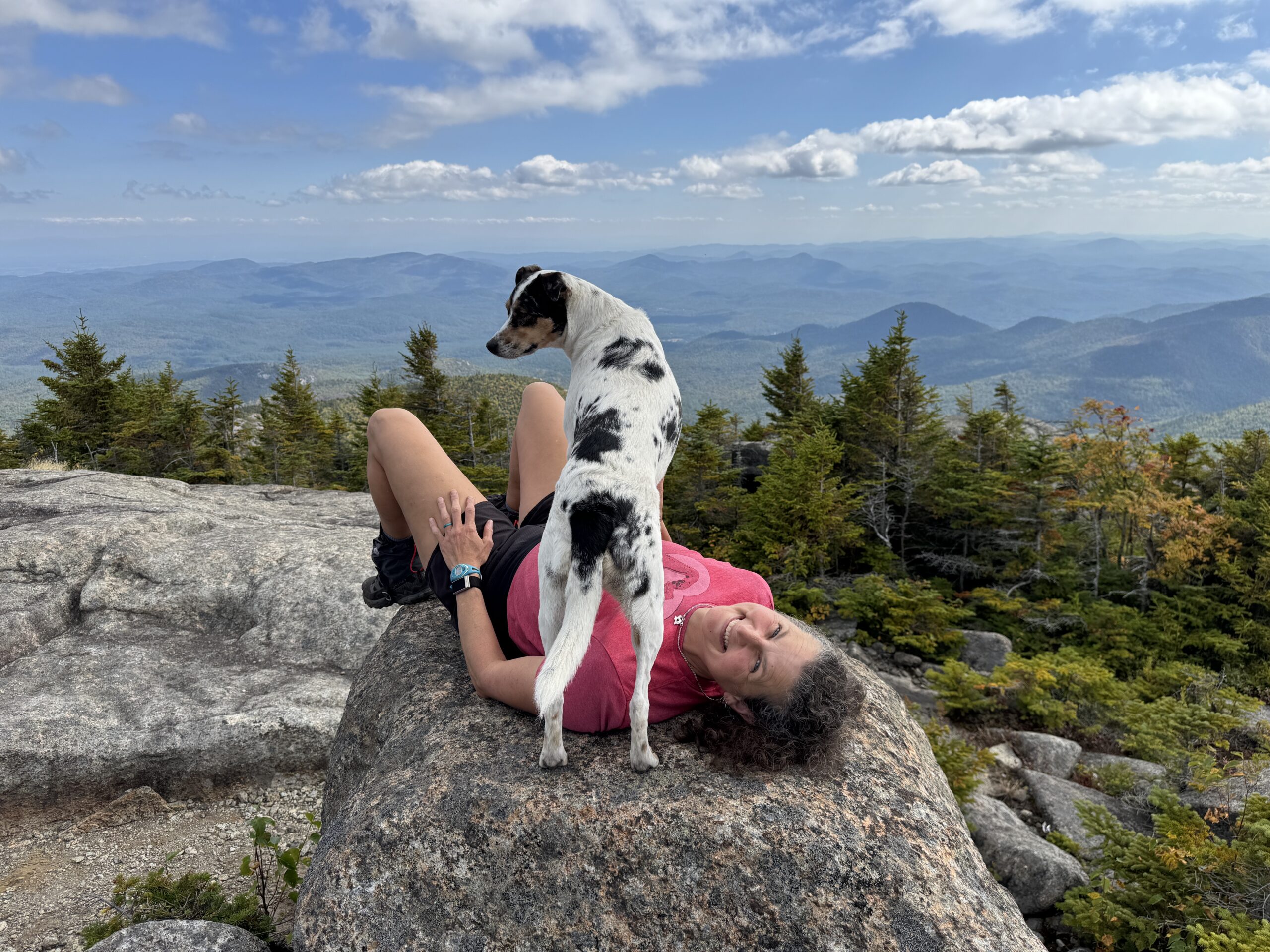 Heidi lays down on a mountain rock with her dog standing over her.